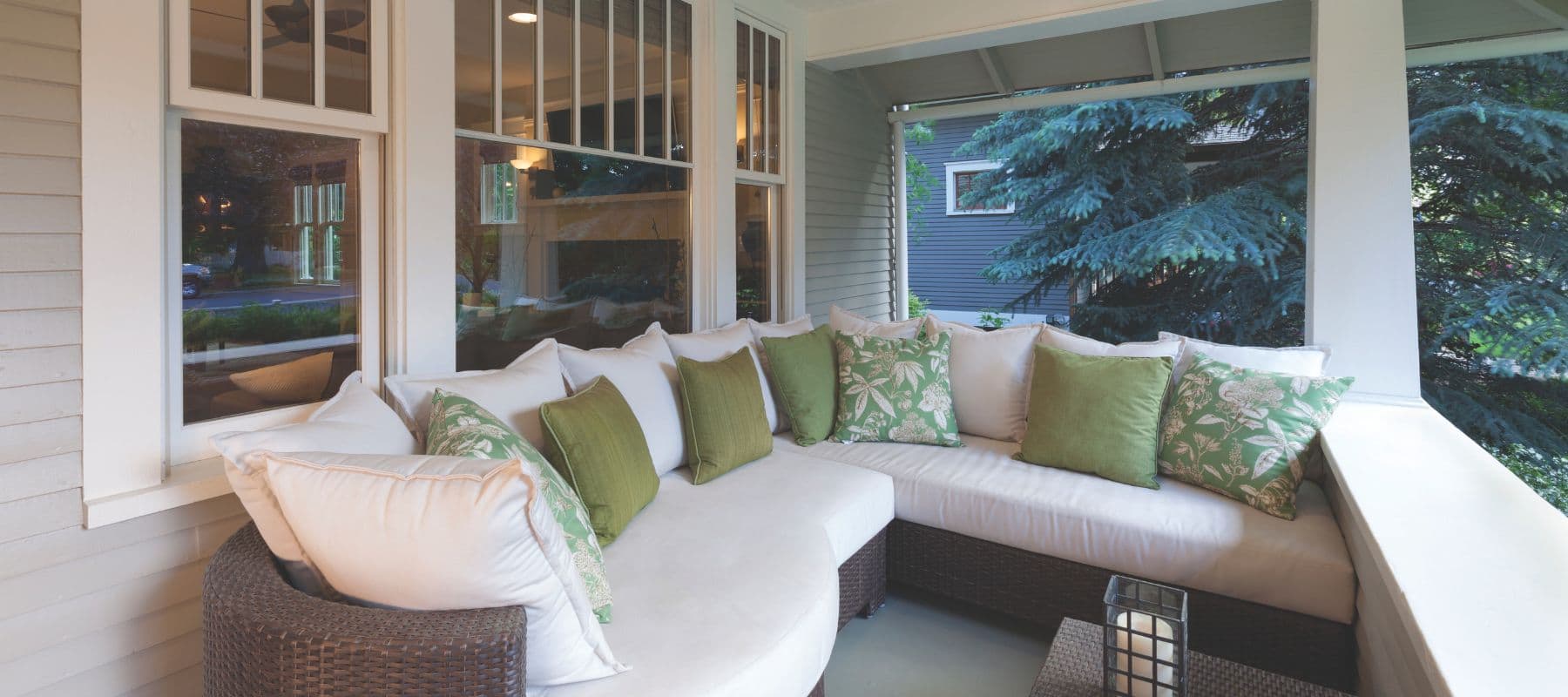 A cozy front porch with a curved wicker sofa, white cushions, and green throw pillows