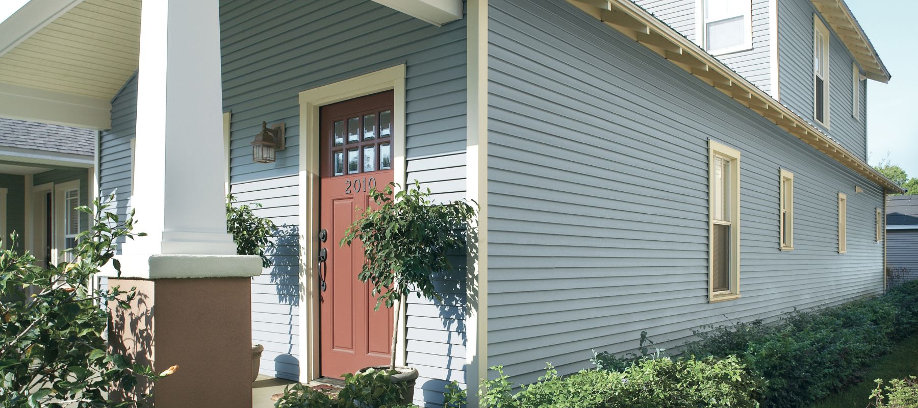 A light blue house with horizontal siding features a reddish-brown front door with windows, white trim, and a prominent front porch column