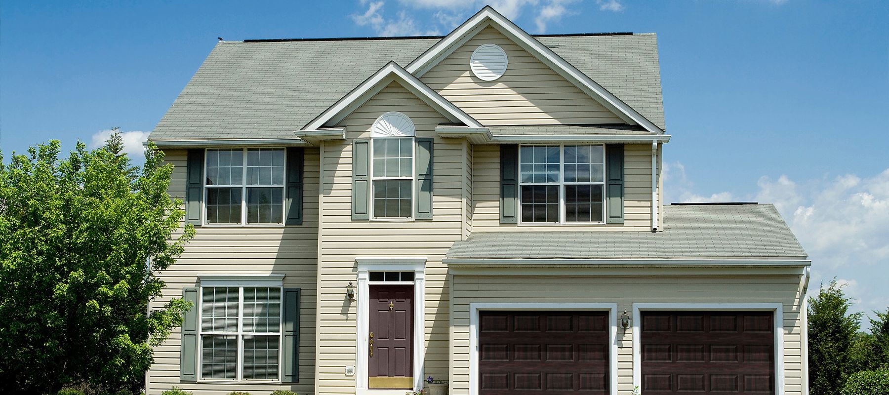 A two-story beige house with gray shutters and a two-car garage stands prominently against a blue sky with white clouds