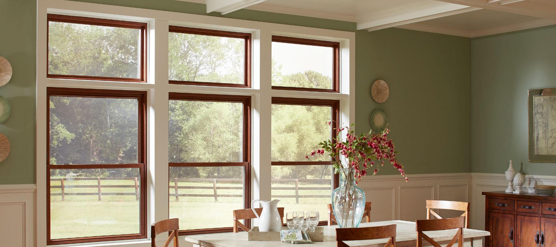 A spacious dining room with green walls and white wainscoting features a prominent set of large brown-framed windows looking out onto a sunny, fenced field and trees